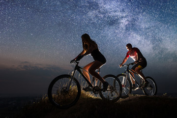 Fototapeta na wymiar Guy and girl riding a bicycles on the hill at night. Starry sky over the two mountain bikers. Bottom view