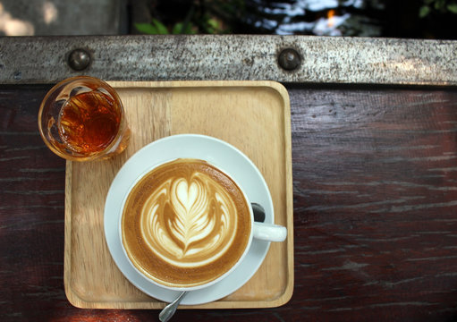 Directly above a hot coffee with froth art served on a wooden tray with a glass of jasmine tea. Rustic wooden bar with industrial bolts and water of the outside setting visible at top of frame