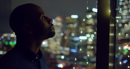 Dark skinned African American man sips from a wine glass in a window with city lights while looking out at Downtown Los Angeles at night. Medium close up, hand held.