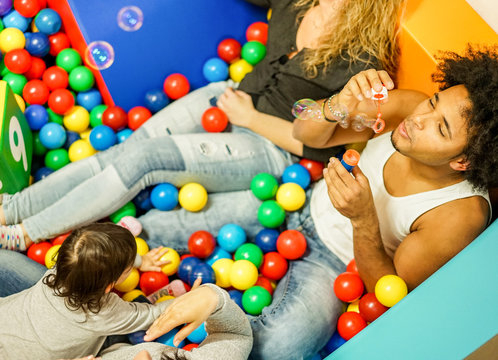 Multi Ethnic Parents Playing With Daughter Inside Ball Pit Swimming Pool - Happy People Having Fun In Children Playground Indoor - Family And Love Concept - Focus On Man Face - Warm Filter