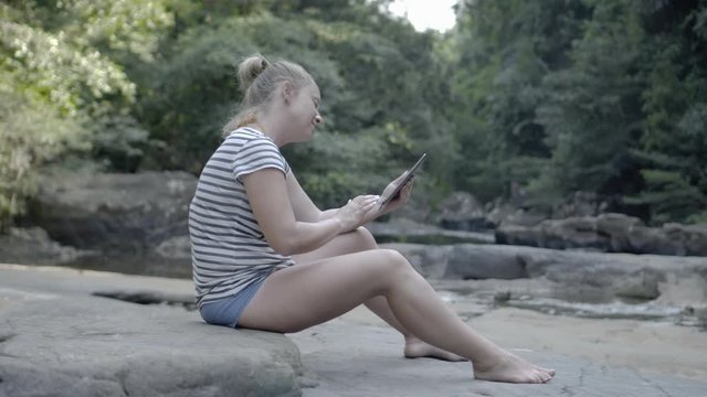 Девушка с планшетом сидит на камне возле горной речки.