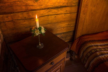 Lit candle in candlestick with white rose decorations, flowers and green leaves standing on rustic night table next to bed in antique, vintage medieval style room. Romantic evening in countryside