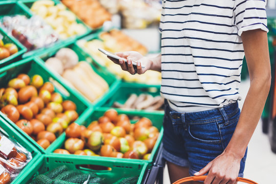 Young woman shopping healthy food in supermarket blur background. Female hands buy products tomato using smartphone in store. Hipster at grocery using smartphone. Person comparing price of produce