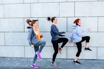 Full height of three sporty women doing a knee-up jump while exercising for marathon and workout fitness near wall.
