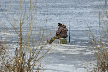 Winter fishing with single man dressed in camouflage sits on a bag in the middle of a frozen river © Vitaly Zubrytsky