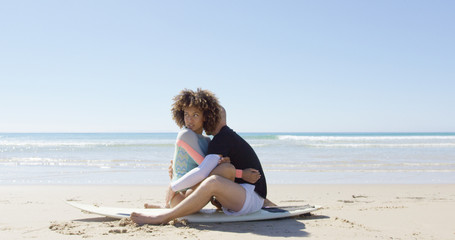 Man and woman sitting on a sunny Tarifa beach and embracing on a surfboard. Horizontal outdoors shot.  © Dash