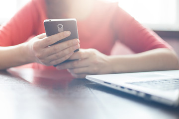 Young woman holding mobile phone and typing messages while sitting at wooden table with open laptop computer. Young woman making online shopping by laptop and smartphone. Modern technology and people.