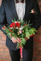elegant man dressed in a modern black suit and white shirt holding in hand a wedding flowers bouquet from red carnation. Close up color vertical image of male hands holding bunch of bright flowers.