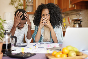 Young dark-skinned family facing financial stress, not able to pay off loan in bank. Depressed African-American man and angry woman working trough papers together, trying hard to make both ends meet