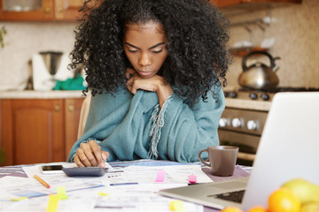 Fototapeta na wymiar Single African-American mother with many debts feeling stressed calculating finances, siting at kitchen table with papers, laptop, making calculations on calculator, trying to make both ends meet