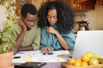 Unhappy and depressed young African-American couple calculating family budget, planning expenses,...