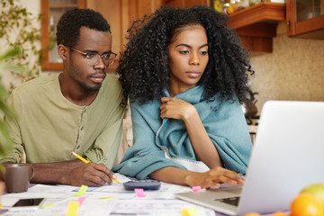 Young African-American family doing paperwork together, sitting at kitchen table with piles of...