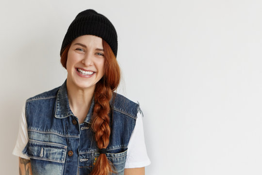 Cheerful student girl in stylish clothes relaxing indoors after college, looking at camera with happy smile. Cute young redhead female with braid wearing black hat and sleeveless jeans jacket at home