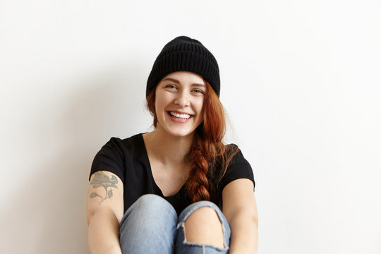 Youth and happiness concept. Indoor shot of joyful young hipster girl with ginger hair in braid and tattoo resting on floor, leaning on white wall, dressed in ragged blue jeans and black hat