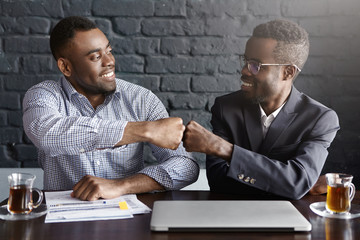 Candid shot of happy successful dark-skinned businessmen wearing formal clothing fist-bumping while...
