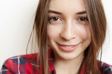 Naklejka premium Highly-detailed close up portrait of beautiful Caucasian teenage girl with dark hair, porcelain skin and brown eyes looking at camera with charming happy smile, dressed in trendy checkered shirt