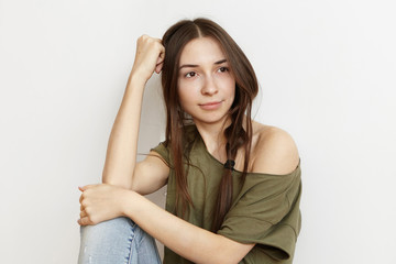 Indoor shot of beautiful young brunette female in stylish clothing, resting elbow on her knee, looking away with thoughtful dreamy expression, dreaming of vacations by sea, siting alone at home