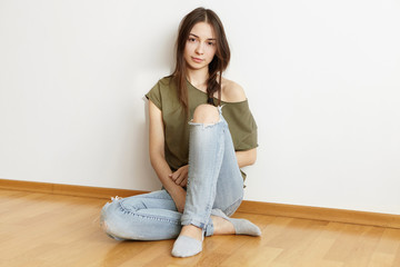 Attractive young brunette woman wearing trendy ragged jeans and top with open-shoulder design sitting on floor at home, leaning on white wall. Pretty girl dressed casually relaxing indoors at weekend