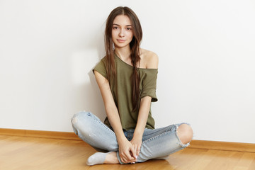 Studio shot of charming woman teenager with messy hairstyle dressed in oversize top and ragged jeans, sitting on wooden floor, keeping her legs crossed. People, leisure and lifestyle concept