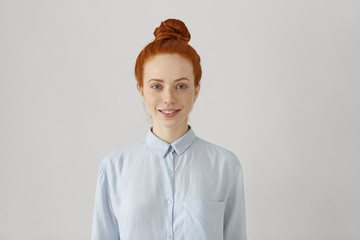 Indoor shot of attractive young redhead female with hair bun in shirt smiling happily, ready for...