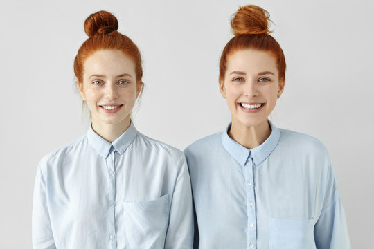 Two young redhead Caucasian females looking alike wearing same formal light-blue shirts, looking at camera, smiling happily, standing close to each other against white studio wall background