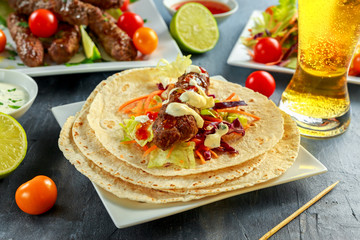 Homemade Kofta kebabs on skewers with pita, lime, vegetables, beer, sweet chili and yogurt sauce on white plate