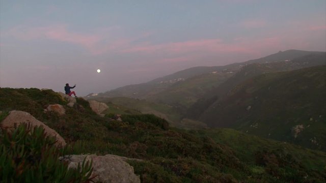 Girl taking photos on the cell phone, sitting on a rocky mountain, beautiful evening moon in the hills, nature sunset sky