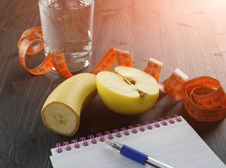 Healthy lifestyle concept - glass of water, apple, banan, measuring tape and notebook on the dark wooden background, toned