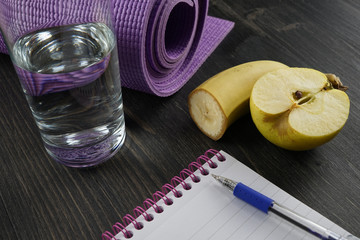 Healthy lifestyle concept - glass of water, fruit, notebook and yoga mat on the wooden dark background