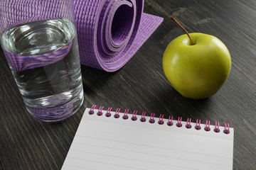 Healthy lifestyle concept - water, apple, notebook and yoga mat on the dark wooden background