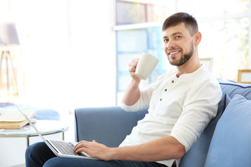 Handsome young man drinking coffee while working with laptop at home