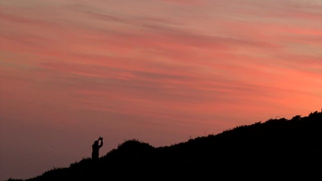 Photographer man silhouette taking photo selfie on the mountain hill, black silhouette on orange sunset sky background