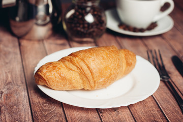 Croissant in a plate, a cup with a drink, a jar with coffee beans and a bucket in the background