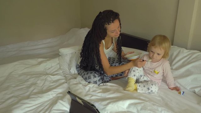 Beautiful young woman feeding little baby girl porridge while looking on tablet computer