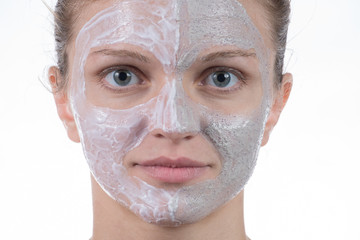 Two cosmetic mask of gray clay with scrub and cream on the face of a young girl on a white background