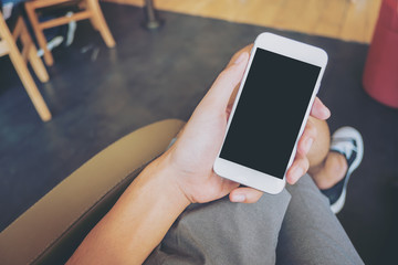 Mockup image of hand holding white mobile phone with blank black screen on thigh with black and white canvas shoes