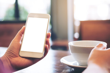 Mockup image of hands holding white mobile phone with blank white screen and hot coffee cup in vintage loft cafe