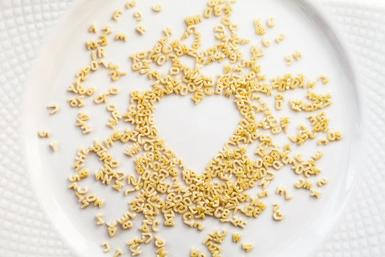 Chaotic alphabet pasta small letters and numerals scattered in a heart shape on a white plate. Uncooked dry food ingredient background. Love for cooking concept