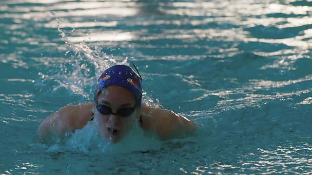 Female swimmer swims in pool HD slow-motion video. Butterfly professional athlete training. Come up from water, splashing, arms stroke. Front view