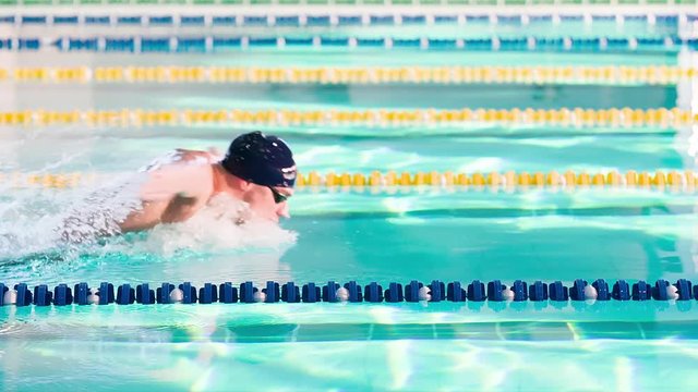 Male swimmer swims in pool HD slow-motion video. Butterfly professional athlete training. Dolphin kick: come up from water, splashing, arms stroke. Side view