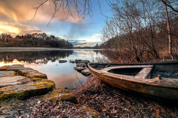 Altes Holzboot am See mit Steinen bei Sonnenuntergang