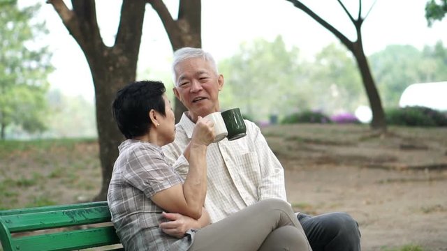 Slow motion happy Asian senior couple sitting and drinking tea, coffee in morning park. Having conversation and showing love