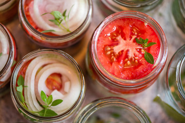 Canning fresh tomatoes with onions for winter in jelly marinade. A shot of basil leaves on top of a red ripe tomato slices and onion rings being put in jar.

