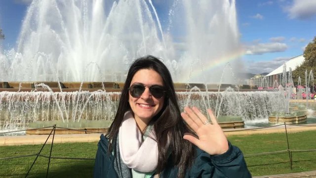 Young woman posing to camera by large funtain and natural rainbow. Girl waving her hand to camera posing for photo
