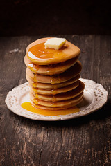 Home made American style pancakes  with golden syrup pored over them and square butter piece on a vintage plate over a rustic wooden board. Dark Rustic Style