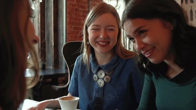 three cute beautiful happy girl smile and laugh, while chatting emotionally in the cafe in the city with cups of coffee during sunny day