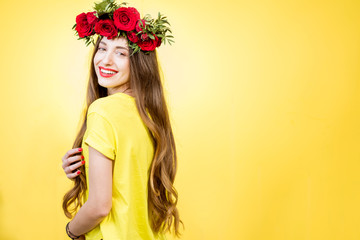 Fototapeta premium Colorful portrait of a beautiful woman in yellow t-shirt with wreath made of red roses on the yellow background