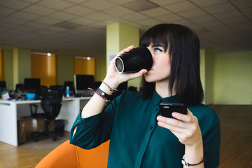 Portrait of a young, beautiful woman office worker that uses the mobile phone and drinking coffee from black cup. Break work. Modern office