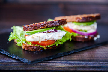 Fresh homemade sandwich with cereals dark rye grain bread, salad lettuce, avocado, tomato, grilled chicken breast, curd cheese and greengrocery on slate stone. Healthy lifestyle breakfast.