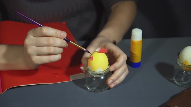 Girl in a dark sweater draws the black paint eyes for a small yellow toy bird. Creating easter chicken from the shell by hand on a grey table close up.